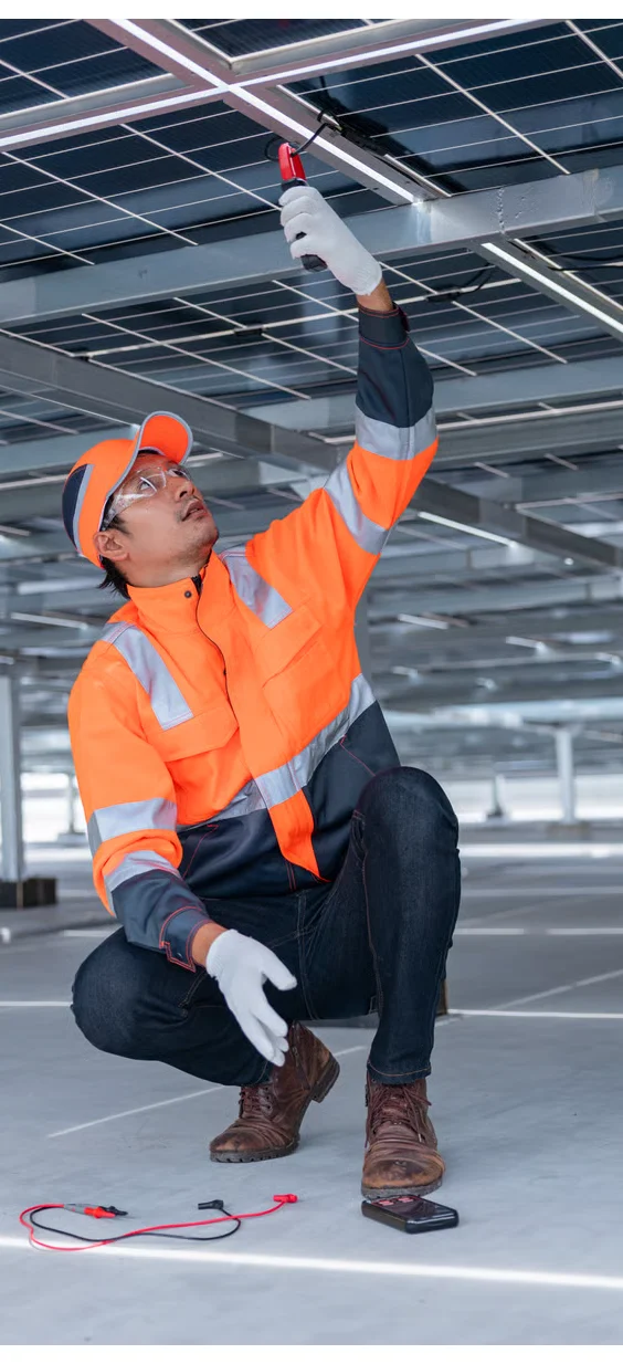 Técnico de mantenimiento revisando paneles solares en un sistema fotovoltaico, utilizando un multímetro para medir voltaje, mientras está vestido con un chaleco de seguridad naranja y guantes.