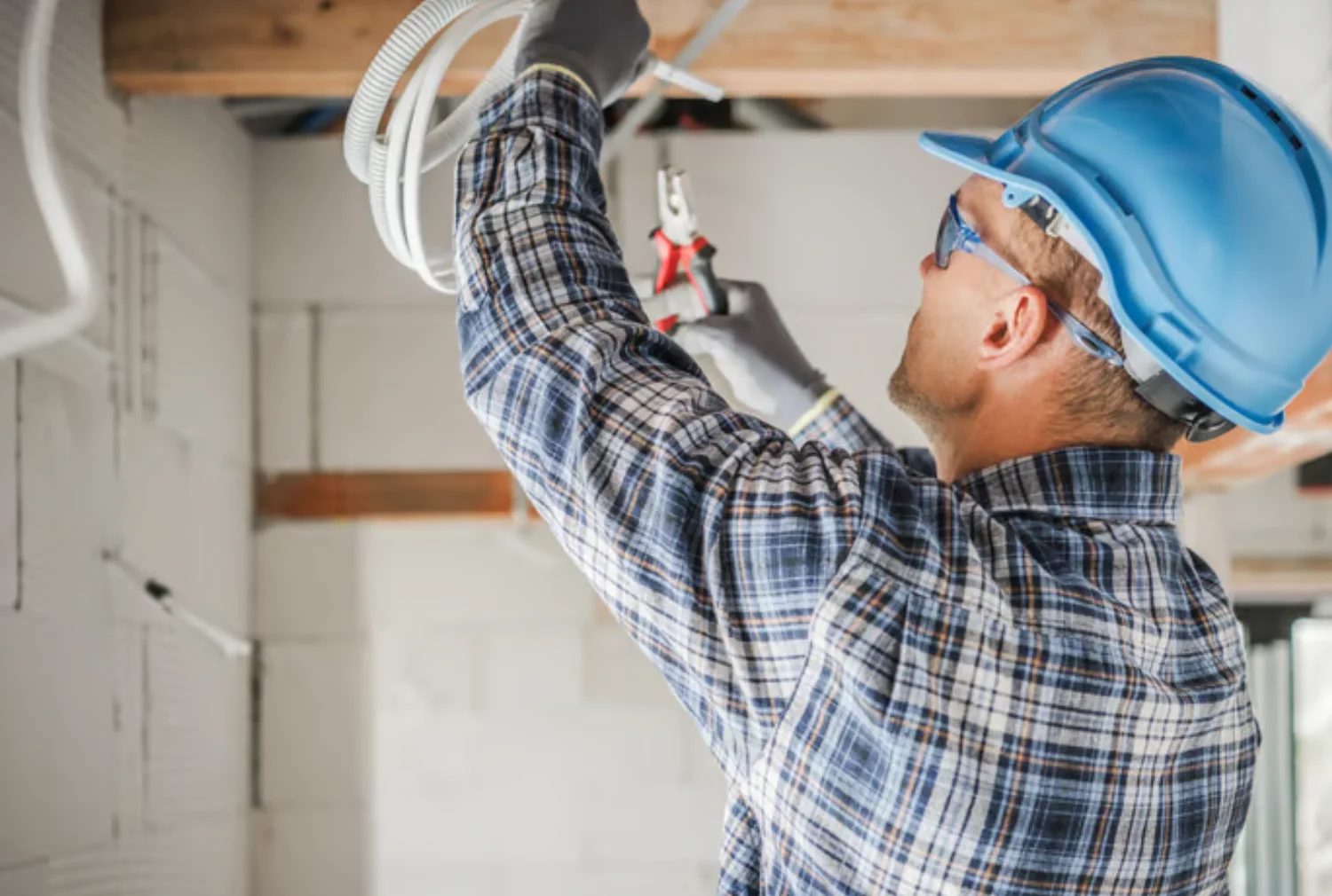 Trabajador de la construcción usando herramientas para instalar cables eléctricos en un techo, llevando casco de seguridad azul y gafas de protección, en un entorno de obra.